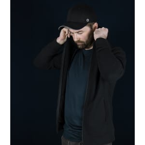 A man in a black Swiss Peak Urban Hoodie adjusts the hood, wearing a black cap. The background is dark.