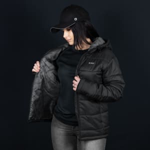 A woman in a black puffer jacket with a hood, wearing a black cap. The jacket features a logo and is partially unzipped.