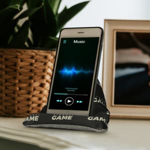 A black bean bag phone holder supports a smartphone displaying a music app, set against a decorative background.