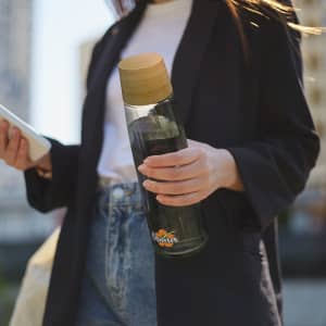 A drink bottle with a glass body and a wooden lid, featuring a logo, held by a person outdoors.