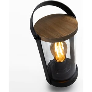 A lantern with a black metal frame, transparent glass, and a wooden top showcasing a glowing light bulb inside.