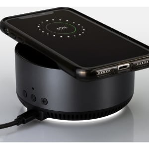 A black wireless charging speaker with a smartphone placed on top, displaying a charge indicator.