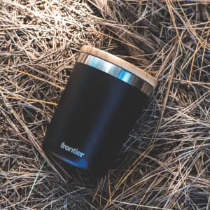 A drink bottle in matte black with a wooden lid, featuring a simple logo on the side, resting on straw.