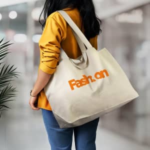 A person wearing a yellow shirt holds a cream tote bag with an orange logo. The background is blurred.