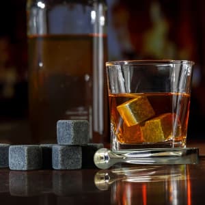 A glass with whiskey and grey whiskey stones beside a drink bottle and a stainless steel tongs.