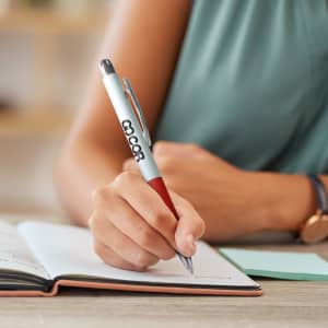 A metal pen in white and red being used to write in a notebook, featuring a logo.