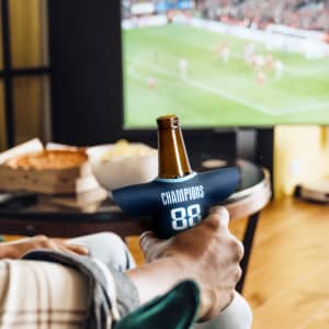 A drink bottle is in a navy blue Jersey Can Cooler with a logo, set next to a TV displaying a sports event.