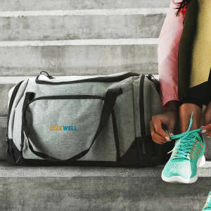 A grey and black duffle bag sits on a concrete surface, with a logo, beside a person tying their shoe.