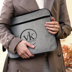 A grey satchel held by a person wearing a trench coat, featuring a logo on the front.