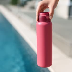 A drink bottle in a vibrant pink colour, being held by a hand near a swimming pool. It features a logo.