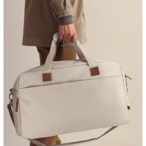 A duffle bag in light grey, being carried by a person with a neutral outfit, featuring a logo and leather accents.