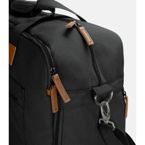 A black duffle bag with tan leather accents and dual zipper pulls, featuring a metal clip and multiple pockets.
