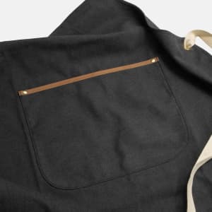 A dark grey canvas apron featuring a back pocket with a tan leather trim and an adjustable strap.