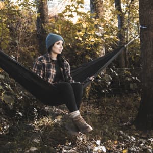 A woman in a beanie and flannel sits in a black outdoor hammock among autumn trees.