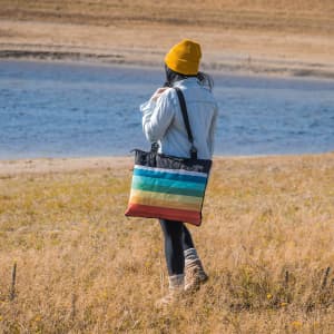 A person wearing a yellow beanie carries a multi-coloured striped picnic blanket against a natural landscape.