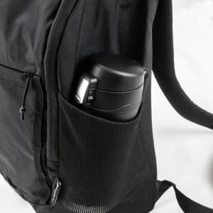 A black daypack features a side mesh pocket holding a black drink bottle. The bag has zippers and adjustable straps.