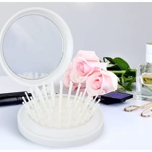 A compact white brush with a mirror, featuring bristles, placed among roses and beauty products.