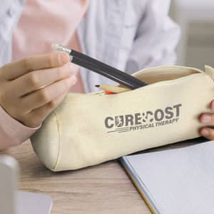 A cream pencil case with a logo, containing pencils and a pen, placed on a wooden surface next to a notebook.