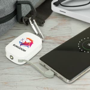 A white power bank with a logo, connected to a smartphone via a grey cable, placed on a wooden surface.