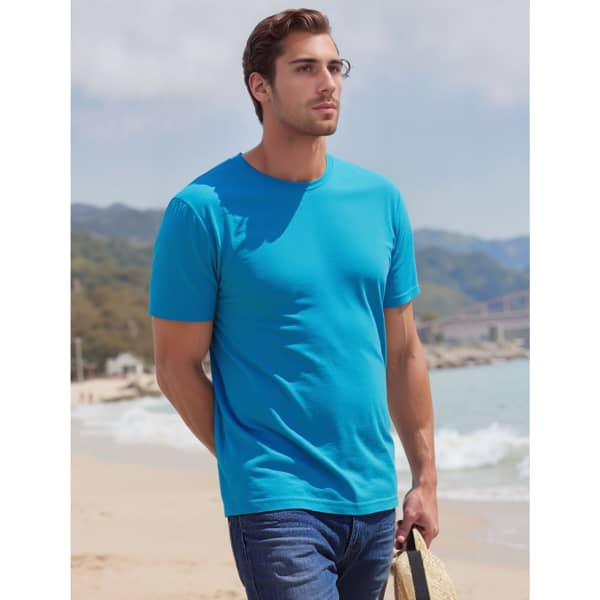 A bright blue t-shirt on a man at the beach, with mountains in the background. The man is holding a straw hat.