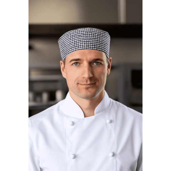 A chef wearing a black and white checkered cap and a white chef's coat, standing in a kitchen environment.