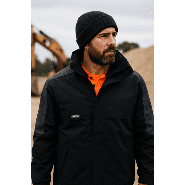 A man wearing a black and charcoal jacket with a beanie stands outdoors, with industrial equipment in the background.