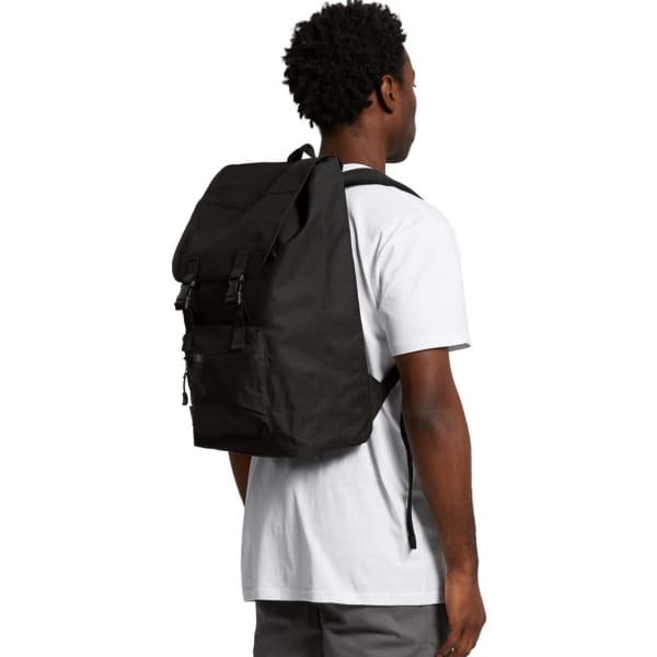 A black recycled field backpack is worn on the back of a person in a white t-shirt. The backpack has adjustable straps and a logo.