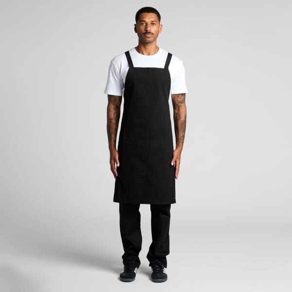 A black apron is displayed on a model, featuring adjustable straps and a simple design, paired with a white t-shirt and black pants.