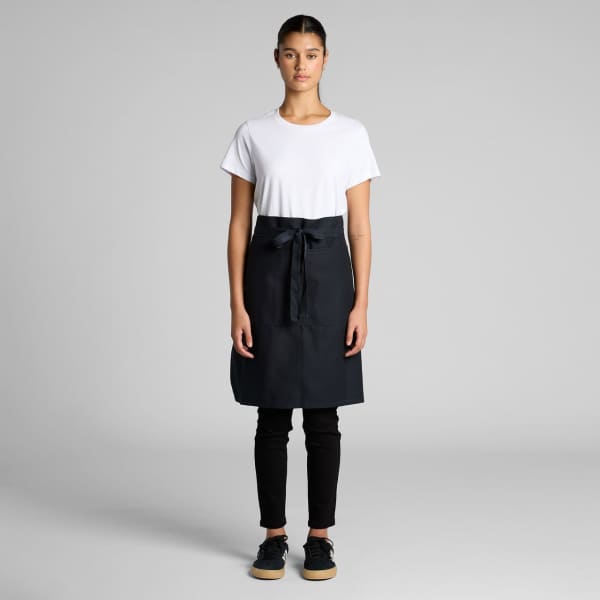 A model wears a black Carrie Half Apron over a white T-shirt and black pants, showcasing the apron design.
