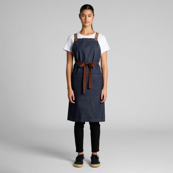 A denim apron in dark blue with brown leather straps and a matching belt, worn by a model against a grey background.