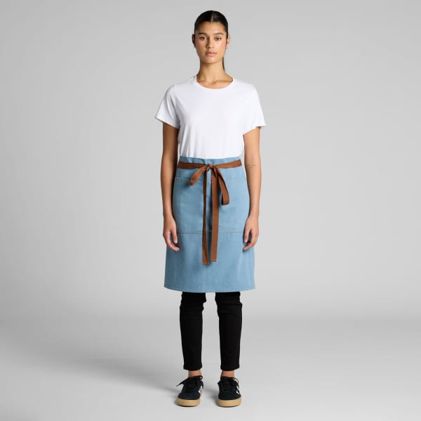 A denim half apron in light blue, featuring a brown waist tie, displayed on a model wearing a white t-shirt and black pants.
