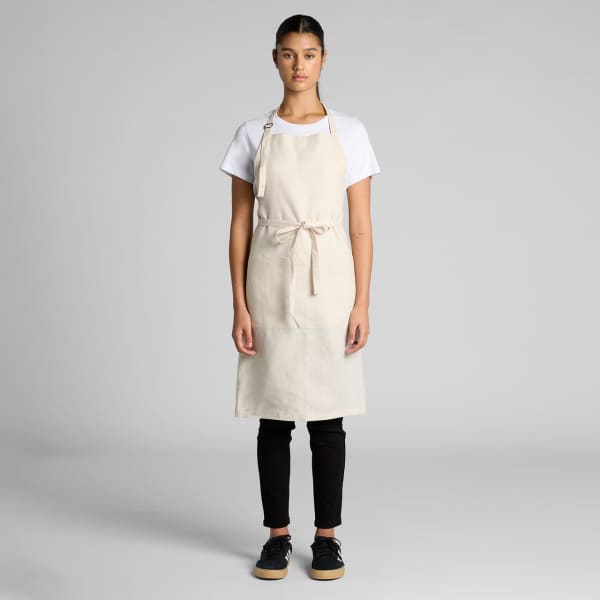 A linen apron in a natural beige colour worn by a model, featuring adjustable straps and a tie waist.
