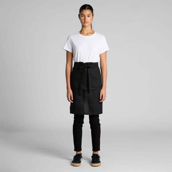 A model wearing a black linen half apron, paired with a white T-shirt and black pants. The apron has a tie at the waist.