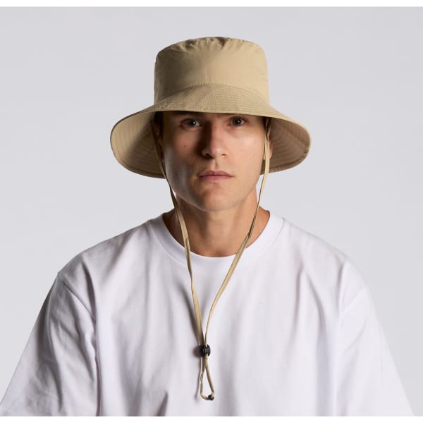 A beige nylon wide brim bucket hat with a drawstring, featuring a logo, worn by a person in a white shirt.