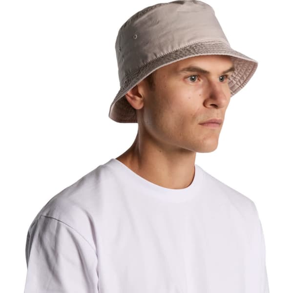 A faded beige bucket hat with a soft brim, worn by a person in a white t-shirt. The hat features a logo.