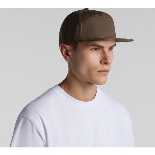 A brown 7-panel cap worn by a model, featuring a logo on the front and a flat brim.
