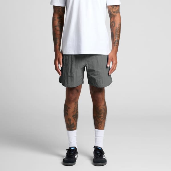 A person stands wearing grey swim shorts, paired with a white t-shirt and black sneakers. The shorts feature a logo.