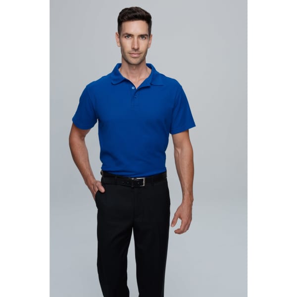 A man stands wearing a blue polo shirt and black trousers, with a logo on the shirt.