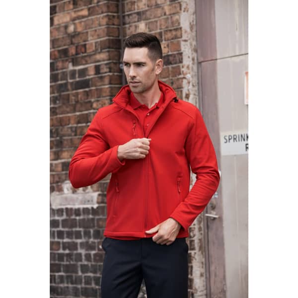 A man in a red soft shell jacket with a hood, featuring zippered pockets, poses against a brick wall backdrop.