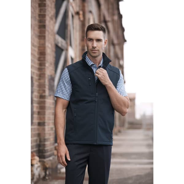 A man in a navy vest over a checked shirt stands confidently in an industrial setting. The vest features a logo.