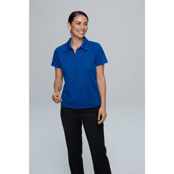 A woman wearing a blue polo shirt and black pants, smiling against a plain background. The shirt features a logo.
