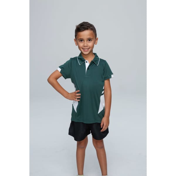 A child in a green polo shirt with white accents stands confidently, smiling, wearing black shorts.