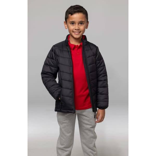A child wearing a black puffer jacket over a red polo shirt and grey pants, smiling while standing.