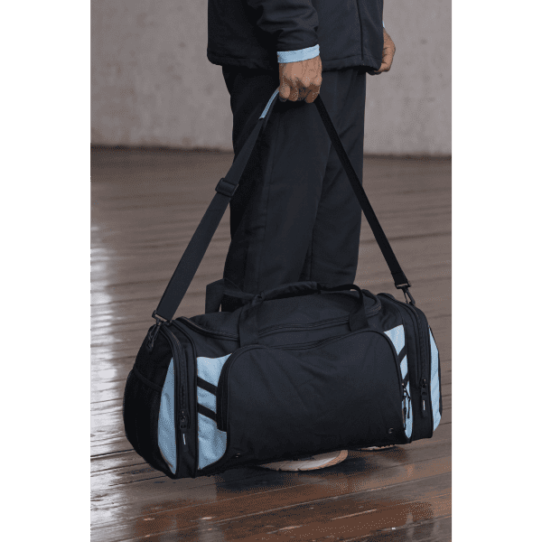 A black and light blue duffle bag being held by a person, featuring multiple zippers and a shoulder strap.