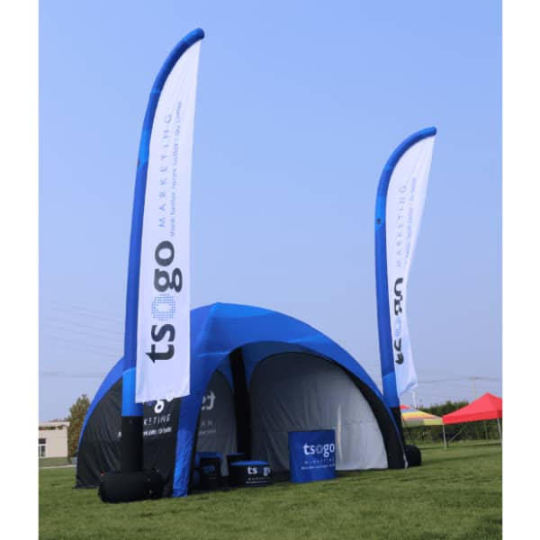 An inflatable tent with blue and white flags featuring a logo, set up on green grass under a clear blue sky.