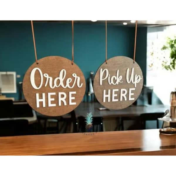 Two round wooden signs hang from a ceiling, one saying "Order Here" and the other "Pick Up Here," in white text.