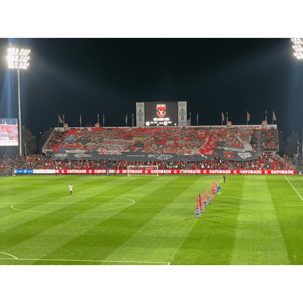 A large polyester TIFO banner in vibrant colours displayed at a stadium, with fans and a field in the foreground.