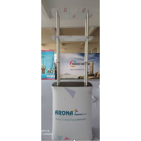 A promotional table in white with shelves, featuring a branding logo, set in a bright indoor space.