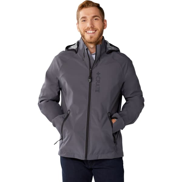 A man wearing a grey Oracle Softshell Jacket, featuring a hood and pockets, stands smiling with hands in pockets.