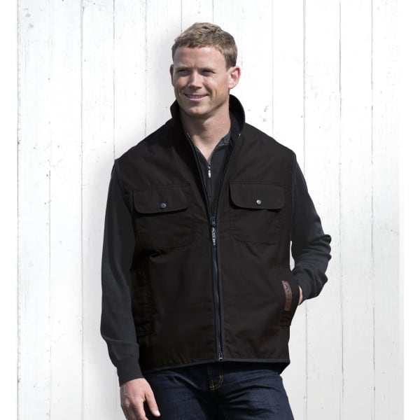 A man wearing a black oilskin vest with two front pockets, standing against a white wooden background.